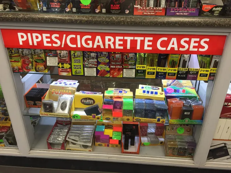 Under our Pipes/Cigarette Cases sign, this case is loaded with flavored cigarillos like 4K's and Game, plus metal and plastic cigarette cases in every color. It's a favorite stop for customers looking for small, affordable tobacco accessories. Glass display case of cigarillos, metal cigarette cases, and pipe accessories at GS Vape in Barberton, Ohio.